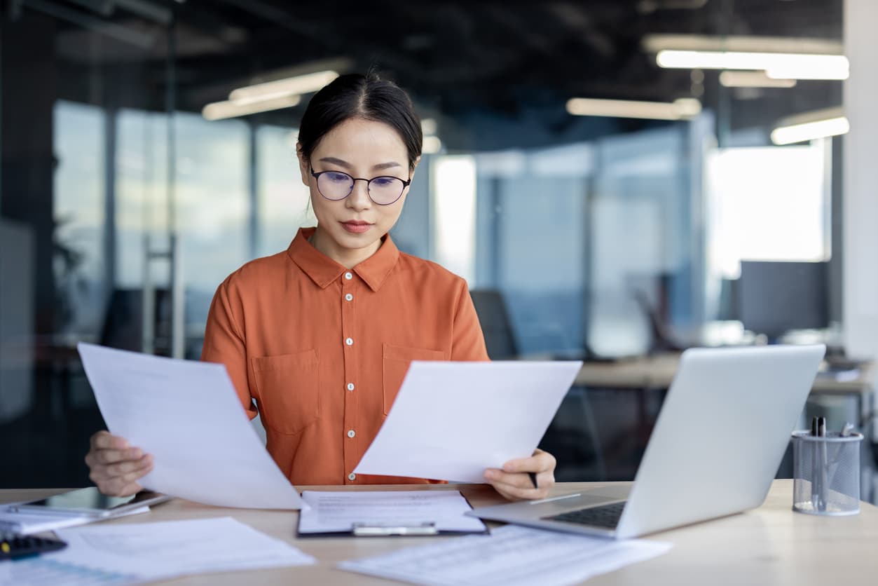 A woman focuses on paperwork with an open laptop in an office.